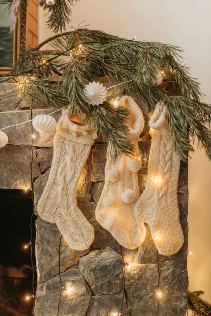 Trois grosses chaussettes tricotés en blanc suspendus à une cheminée décorée de branches de conifères et de lumières féériques.
