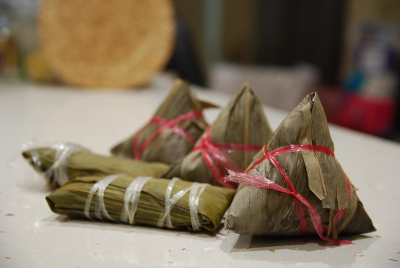 Cinq zongzi, boules de riz gluant emballés dans une feuille et attachés par une ficelle. Trois sont triangulaires, deux sont cylindriques.