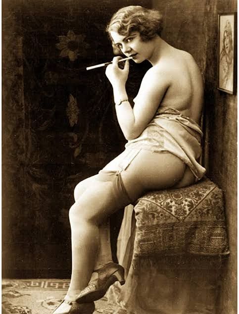 Photo érotique des années 1920, montrant une femme assise sur un coussin, légèrement vêtue, tenant une cigarette à la main. Elle pose dans un cadre vintage, avec un regard suggestif.