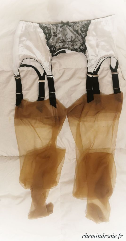 Un porte-jarretelles blanc avec de la dentelle noire et des bas transparents de couleur marron, posés sur une surface plane.