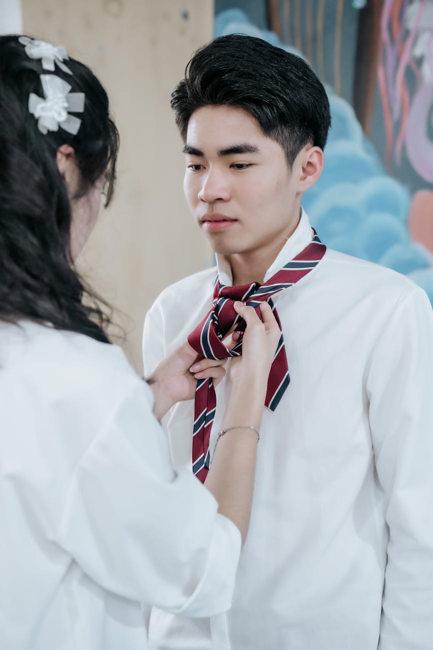 Une femme nouant la cravate d'un jeune homme asiatique. Ils sont tous les deux vêtus de blanc, la cravate est rouge à rayures diagonales bleues.